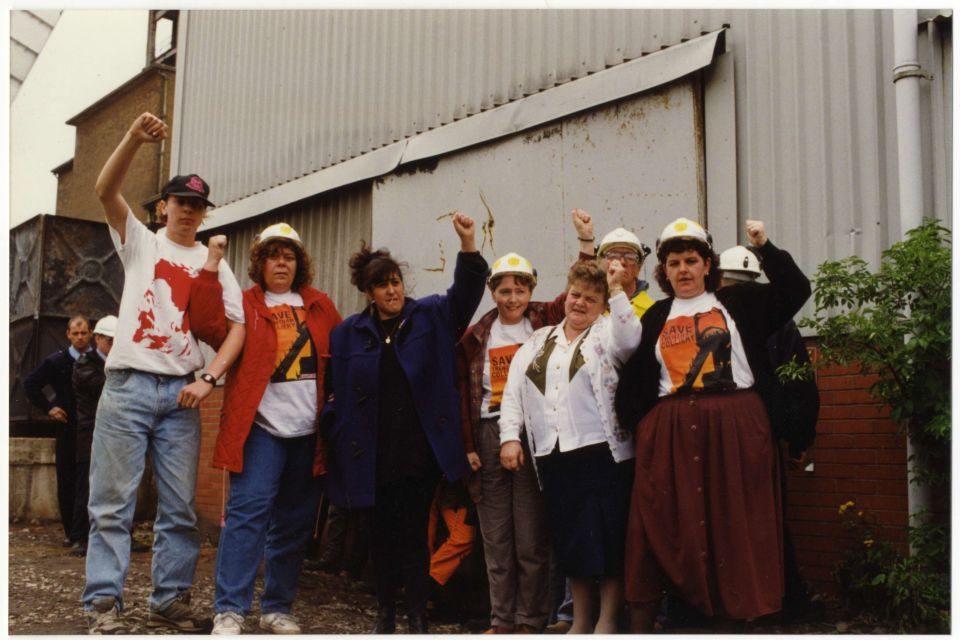 A group of eight individuals, six women and two men, stand together with fists raised in solidarity. Five are wearing hard hats, including the Trentham 3: Brenda Procter, Bridget Bell, and Gina Earl, who are also wearing T-shirts that read 'Save Trentham Colliery.' In the background, two men, possibly security personnel or police officers, look on